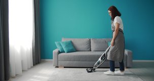 Full length portrait of mature woman in domestic outfit removing dust from white carpet with vacuum cleaner. Housewife in grey apron cleaning own apartment.