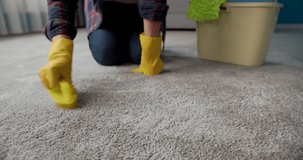 Mature housewife in domestic outfit and rubber gloves standing on knees while cleaning carpet with brush, close up. Concept of chores.