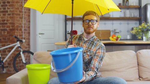 Portrait of upset young hipster guy in glasses sitting on couch at home under umbrella holding plastic bucket collecting water falling from ceiling