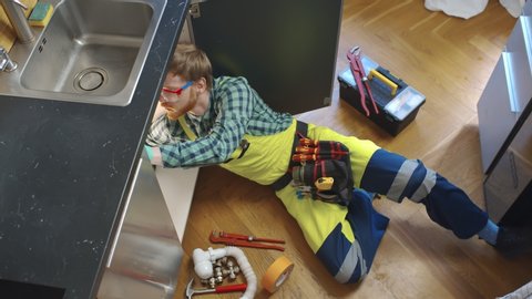 Top view of young plumber lying on floor fixing broken sink pipes in kitchen with tools around. High Angle View Of Male Plumber In Overall Fixing Sink Pipe