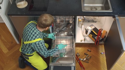 Top view of young handyman installing shelves in dishwashing machine. Handsome repairman fixing broken dishwasher in modern kitchen