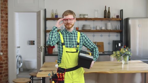 Handyman holding tools standing in kitchen and putting on protective glasses smiling in camera. Portrait of cheerful plumber in workwear with equipment