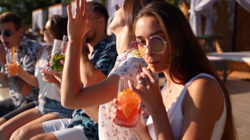 Pretty woman in stylish sunglasses toasting with friends, drinking cocktail of plastic straw at pool party. Group of friends having fun clinking glasses with beverage sitting at poolside. Slow motion. | Shutterstock HD Video #1054066838