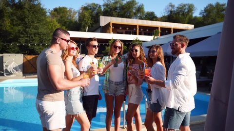 Group of friends having fun at poolside summer party clinking glasses with summer cocktails on sunny day near swimming pool. People toast drinking fresh juice at luxury tropical villa in slow motion.