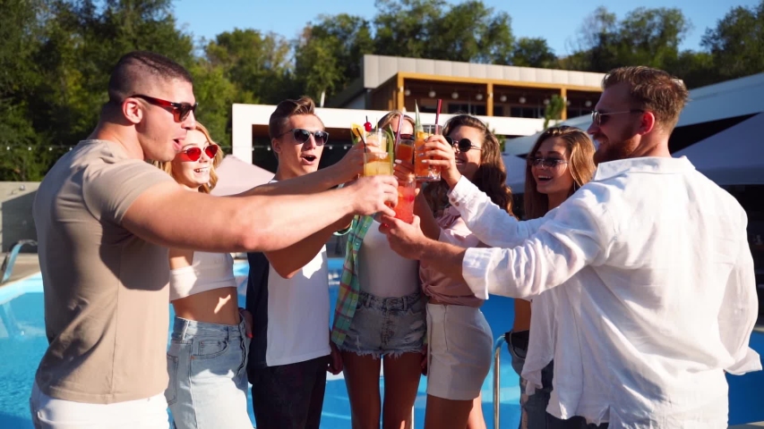 Group of friends having fun at poolside summer party clinking glasses with summer cocktails on sunny day near swimming pool. People toast drinking fresh juice at luxury tropical villa in slow motion. | Shutterstock HD Video #1054066847