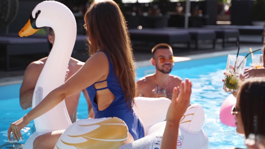 Friends having party with cocktails in holiday villa swimming pool. Happy young people in swimwear dancing, clubbing with inflatable flamingo, swan, mattress in luxury resort on sunny day. Slow motion | Shutterstock HD Video #1054066859
