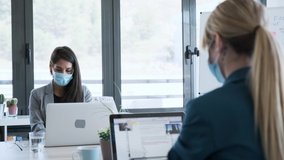 Video of two business women wearing a hygienic face mask while work with laptops in the coworking space. Social distancing concept.