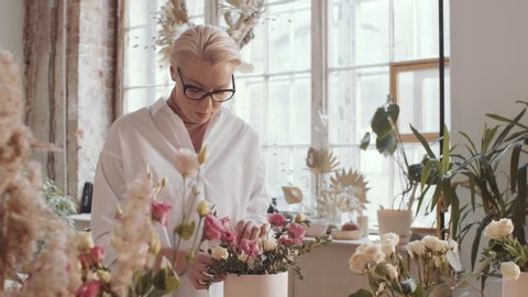 Beautiful middle aged female florist in glasses arranging roses composition and looking at camera with happy smile while working in flower shop