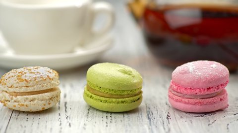 Three sweet macaroon cakes on the table. White and orange, green, pink cakes. Teapot and a cup in background. Slider shot, 4K