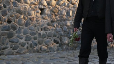 Two people dressed in black walk together down a cobblestone path while solemnly holding flowers. Slow motion waist height shot.