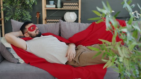 Portrait of guy in super hero costume red cape and mask sleeping on couch at home relaxing during day time. People, lifestyle and relaxation concept.