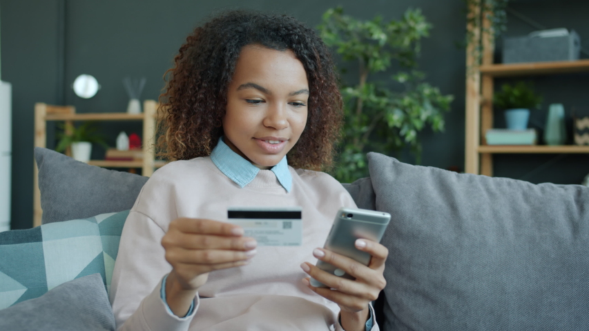 Joyful African-American girl is shopping online with bank card and smartphone from home touching screen holding plastic card and smiling enjoying activity. Royalty-Free Stock Footage #1054075838