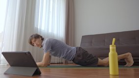 A young man at home doing physical exercises following instructions from a video he watches on a tablet. Social distancing concept. Internet trainer concept