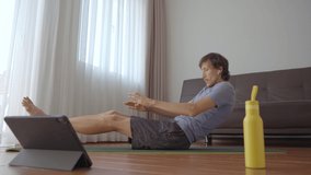 A young man at home doing physical exercises following instructions from a video he watches on a tablet. Social distancing concept. Internet trainer concept