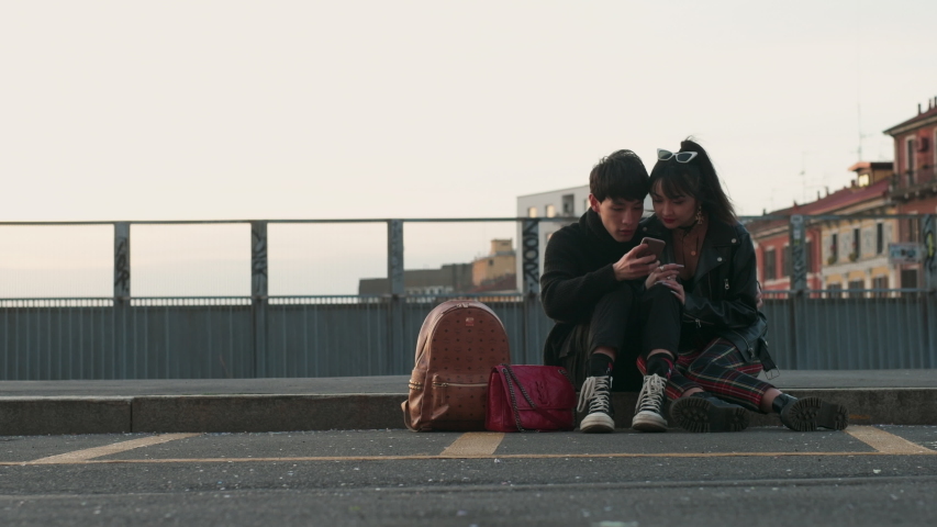A young man and woman seated on the sidewalk at dusk, sharing a phone, both looking at the screen.