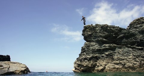 A man wearing a wetsuit diving off a high cliff into the sea.