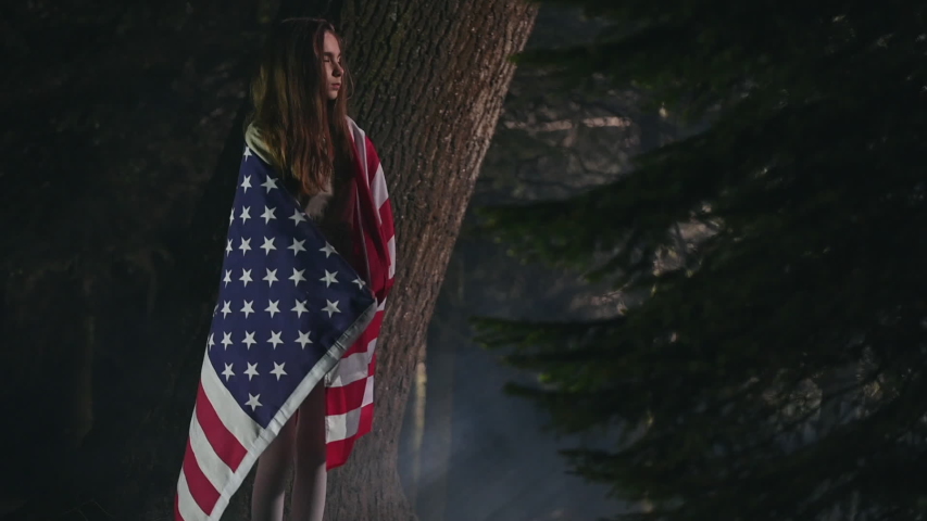 School Age Girl Standing Among Trees With Big American Flag Wrapped Around Shoulders. Child Opens Her Arms Holding And Displaying Flag.