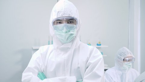 Asian man scientist is wearing Personal Protective Equipment (PPE) looking at camera while standing with his arms crossed and Showing thumb up in a laboratory 
