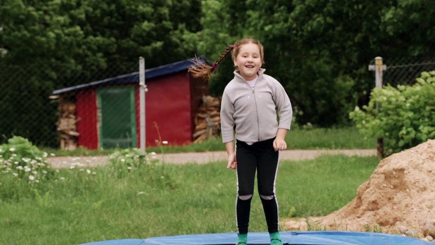 A young girl of preschool age jumps on a trampoline. She smiles and laughs. A trampoline stands on the street in the courtyard of the house