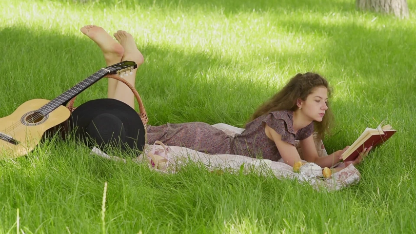 a young girl in a summer sundress lies on a blanket and reads a book in nature