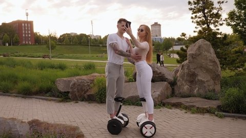 A beautiful girl in white pants rides with a guy on gyro scooters or hoverboards in a summer park, they take a selfie on the phone. Slow motion.