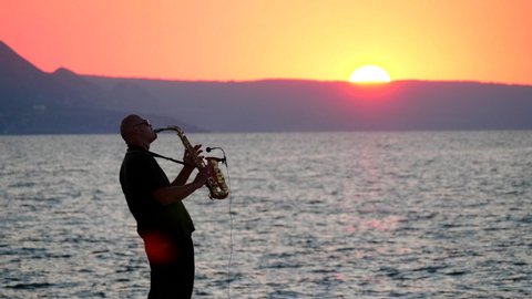 Silhouette of young male saxophonist musician playing golden alt saxophone on musical instrument on seashore, amid waves, beautiful orange sunset, big sun, dawn. Records melody into microphone.