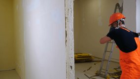 Worker in uniform and respirator destroys inner wall sledge hammer. Demolition work and rearrangement concept. Wall made of gypsum cardboard being destroyed. 4 k video