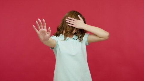 Don't want to watch! Displeased young brunette woman in polo t-shirt covering eyes and gesturing stop, confused and shy to look at shameful content. indoor studio shot isolated on red background