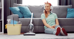Happy blonde woman listening music in wireless headphones while relaxing on floor after spring cleaning. Concept of housework and enjoyment.