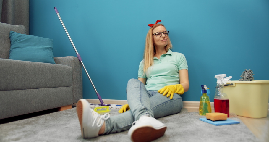 Attractive lady in glasses and rubber gloves sitting on floor with all cleaning products. Mature woman feeling tired of cleaning own apartment.