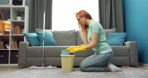 Side view of mature woman with blond hair sitting on knees near bucket with cleaning tools and products. Competent housewife in rubber gloves preparing for removing dust on carpet.
