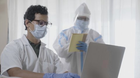 Two men doctors or male healthcare workers wearing an apron and personal protective kit working on a laptop together in the hospital or clinic amid Coronavirus or COVID 19 epidemic or pandemic