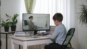 school online, boy learns using video lessons with teacher, looks at the monitor screen and answers tasks while sitting at home at table with books during isolation and quarantin