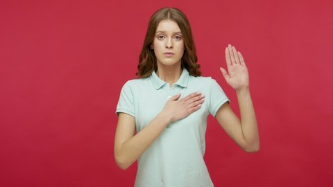 I swear to be honest! Sincere responsible young brunette woman in polo t-shirt raising hand to promise, taking vow with serious dedicated expression. indoor studio shot isolated on red background