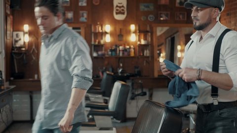 Stylishly dressed Barber dancing in a barbershop.
Brutal Barber with a beard wipes the hair of the client before cutting. Dancing Barber.