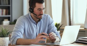 Focused young businessman in eyeglasses holding video conference call with clients, wearing wireless headphones with microphone. Skilled happy salesman advertising goods to customers, writing notes.