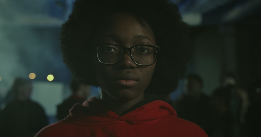 Close up view of young african american girl in glasses looking to camera while standing at demonstration at night street. Crowd of protesters at background. Concept of strike. Black Lives Matter. Royalty-Free Stock Footage #1054134623
