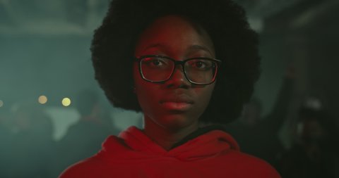Close up view of young african american girl in glasses looking to camera while standing at demonstration at night street. Crowd of protesters at background. Concept of strike. Black Lives Matter.