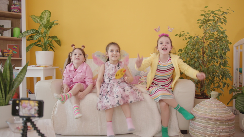 Shooting a Video Blog for Children with Smartphone. Production of Kid's Content. Three Cute Cheerful Little Caucasian Girl Dancing and Laughing, Cozy Interior with Yellow Wall on the Background. Royalty-Free Stock Footage #1054135160