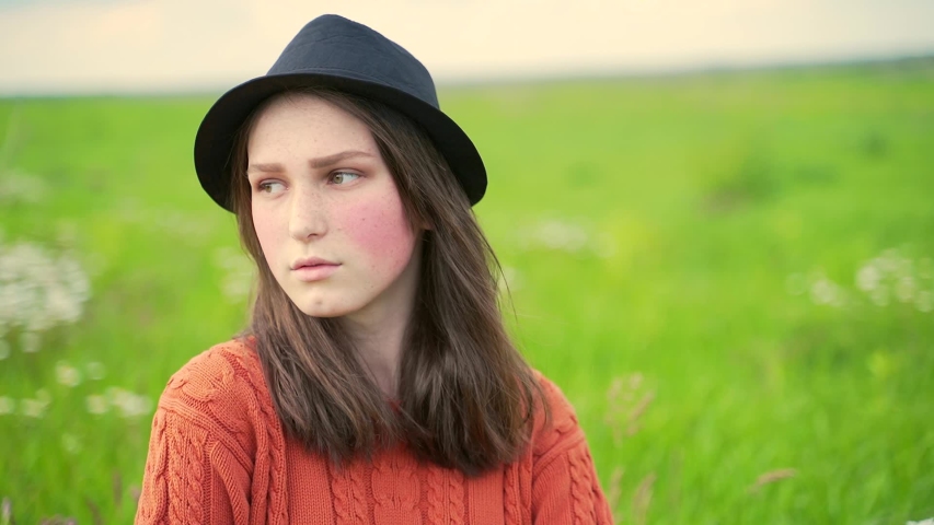 
Portrait of a young rural girl in a hat and sweater sitting on the lawn on the grass. Teen girl with freckles and red cheeks in nature with close up. Green eyes and a hole in the chin