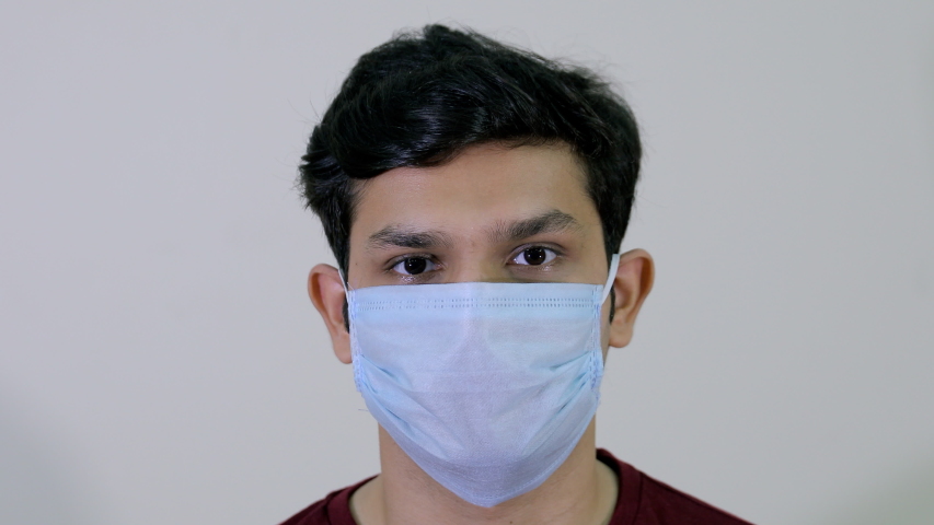 A young attractive boy removes his protective medical mask to breathe fresh air. Closeup of an Indian guy takes off his surgical mask during the Covid-19 pandemic against the white background Royalty-Free Stock Footage #1054138925