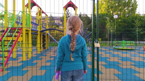 Sad lonely child near the closed playground because of the quarantine.A girl with long blonde braided hair staying alone near empty fenced showground without children.COVID-19 lockdown social distance