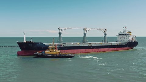 Aerial drone video of cargo container ship guided by yellow tug assistive boat to logistics commercial port. Tugboat transports a large ship to the port dock. Drone shoot.