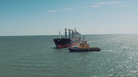 Aerial drone video of yellow tugboat goes to a large cargo ship to transport to the dock. Cargo ship barge and tugboat sail to meet each other in the seaport of the port. Aerial View. Drone shoot.