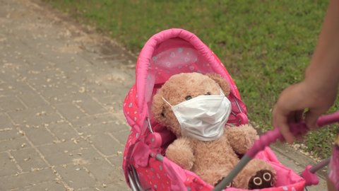 child plays an epidemic and protects toy. girl on street with his favorite toy in protective mask. healthy childhood concept. little girl walks in park with a pram and a teddy bear in medical mask.