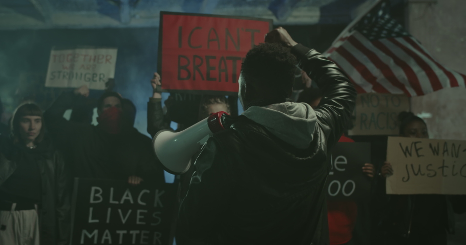 Afro american guy with megaphone turning to camera and walking while raising clenched fist up and shouting with protesters crowd behind. Strike against police brutality and racism.Black Lives Matter. Royalty-Free Stock Footage #1054144532