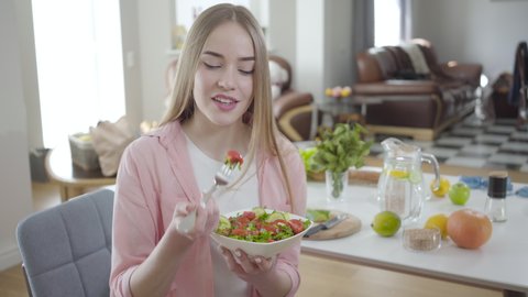 Positive young woman chewing tasty organic salad and smiling at camera. Portrait of joyful blond lady eating healthful vegetable dish. Healthy lifestyle, weight control, beauty.