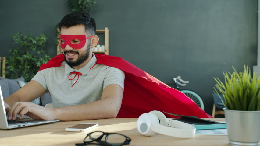 Portrait of young man in superhero costume working with laptop in apartment typing sitting at desk alone and smiling. Modern technology and occupation concept. Royalty-Free Stock Footage #1054148429