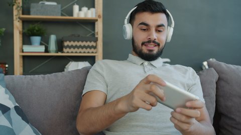 Joyful Arab man is enjoying music through wireless headphones and playing video game in smartphone sitting on sofa in apartment. People and gadgets concept.
