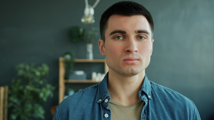 Slow motion of handsome guy with serious face smiling standing alone indoors in apartment and looking at camera changing facial expression. Human emotions and people concept. | Shutterstock HD Video #1054148588